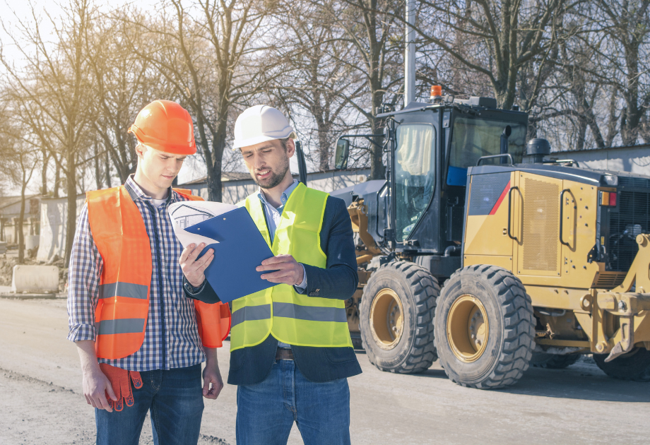 Two construction professionals in hard hats and safety vests reviewing project documents at a job site beside heavy construction equipment during a site inspection.